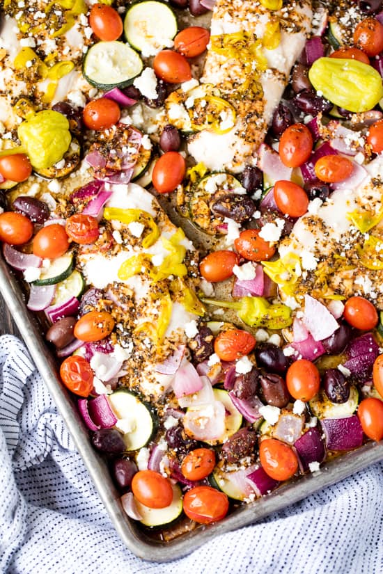 Sheet Pan Greek Chicken and Veggie Bake with cherry tomatoes, olives, red onion, zucchini, pepperoncini and topped with crumbled feta cheese on a sheet pan