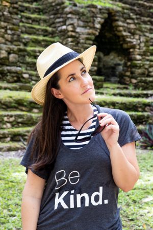 Rachel looks pensively with her sunglasses resting on her chin in front of a Mayan ruin. Her tshirt says "Be Kind."