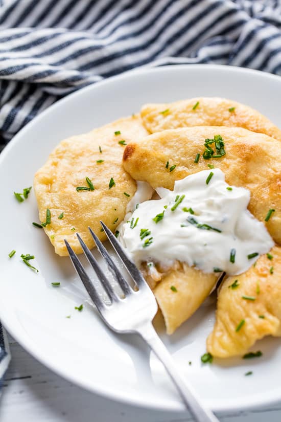 A plate of Potato and Cheese Pierogis with sour cream and topped with freshly chopped chives with a bite taken out of one