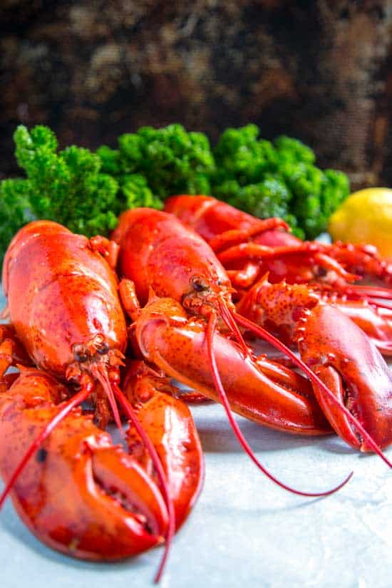 Three cooked lobsters on a serving plater with leafy greens and lemon in the background.