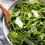 Arugula salad in a white bowl with a wooden spoon in it.