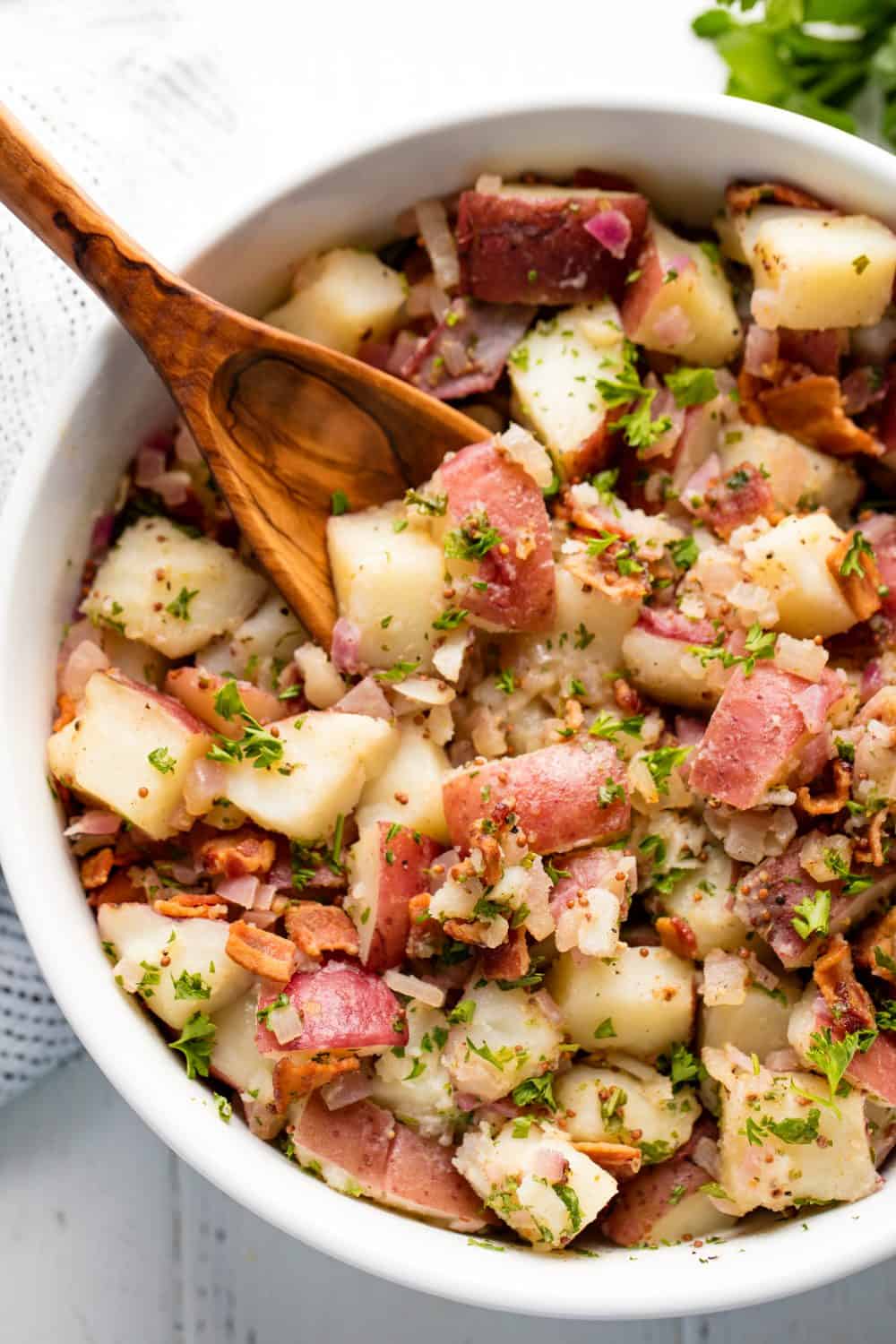 German Potato Salad in a white bowl with a wooden spoon in it.