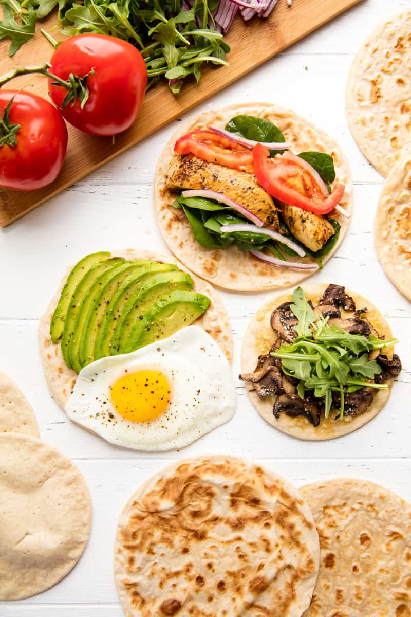 Bird's eye view of 3 flatbread recipes, an avocado and egg flatbread, a muhsroomm and arugula flatbread, and spinach, onion, grilled chicken, and tomato flatbread.