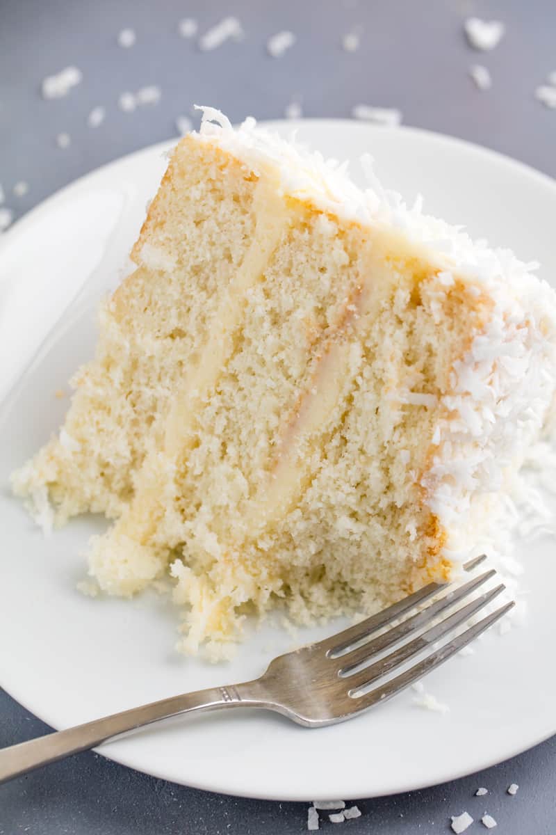 A slice of Coconut Cake on a white plate with a fork and coconut sprinkled around the plate
