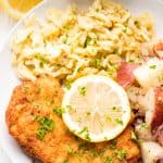 Schnitzel, spaetzle, and german potato salad on a white plate topped with lemon and minced parsley.
