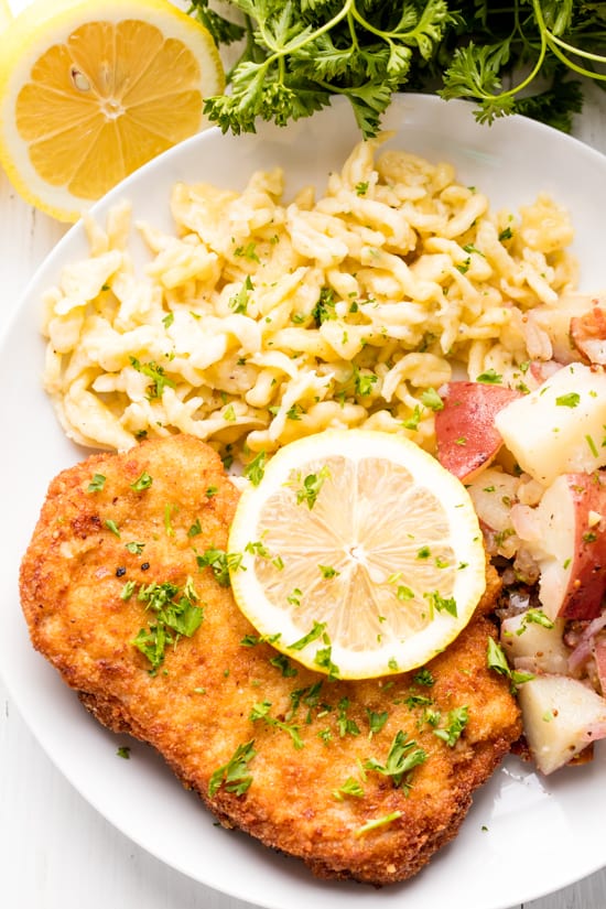 Schnitzel, spaetzle, and german potato salad on a white plate topped with lemon and minced parsley.