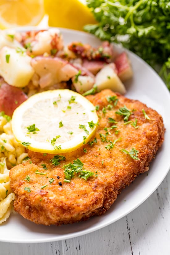 Schnitzel on a white plate topped with a sliced lemon and minced parsley. 