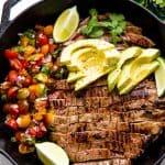 Mexican Skillet Steak in a skillet topped with avocado slices and two lime wedges all sitting next to a side of pico de gallo.