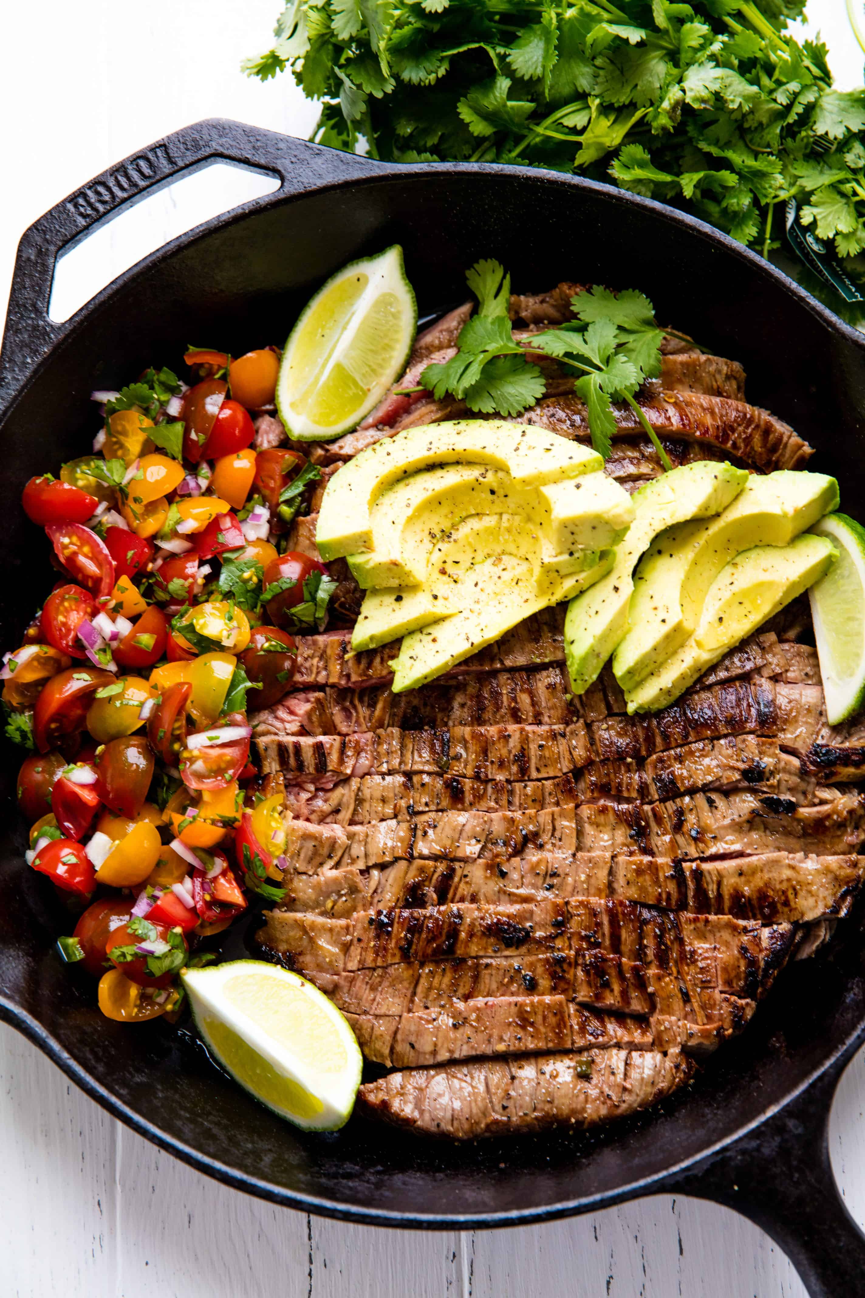 Mexican Skillet Steak in a skillet topped with avocado slices and two lime wedges all sitting next to a side of pico de gallo.
