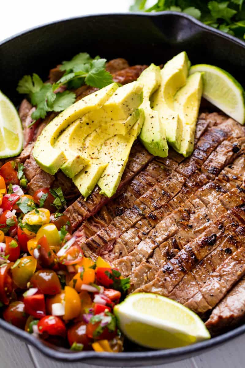 Close up of Mexican Skillet Steak in a skillet topped with avocado slices and two lime wedges all sitting next to a side of pico de gallo.