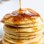 Stack of eight pancakes with a pat of butter getting syrup drizzled on it.