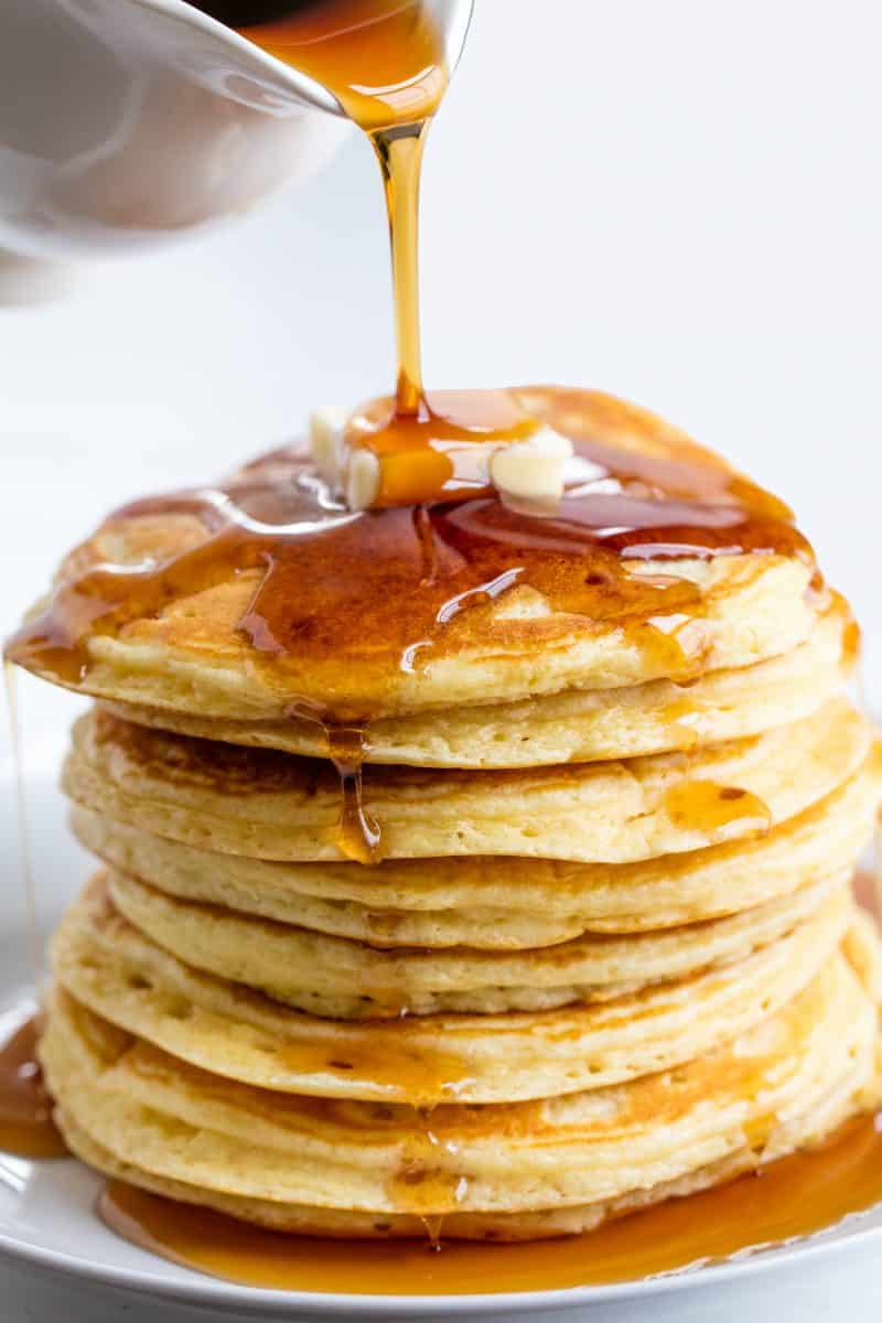 Stack of eight pancakes with a pat of butter getting syrup drizzled on it on a white plate..
