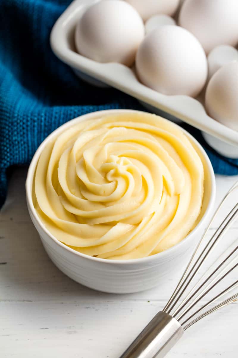  Bird's eye view of pastry cream swirled into a white bowl with a whisk in front of the bowl and eggs behind the bowl. 