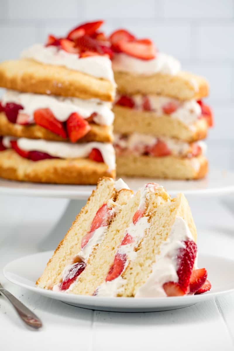 A slice of Strawberry Shortcake on a white plate with the rest of the cake behind it.