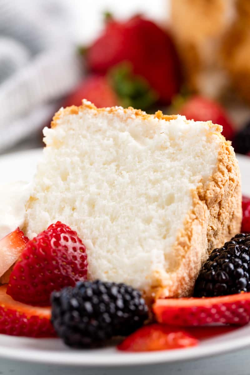 Slice of angel food cake on a white plate with strawberry slices and blackberries. 