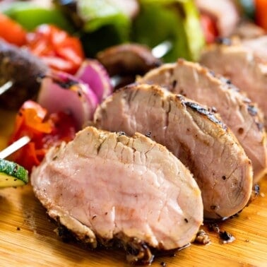 Slices of grilled pork tenderloin by grilled vegetables on a cutting board.