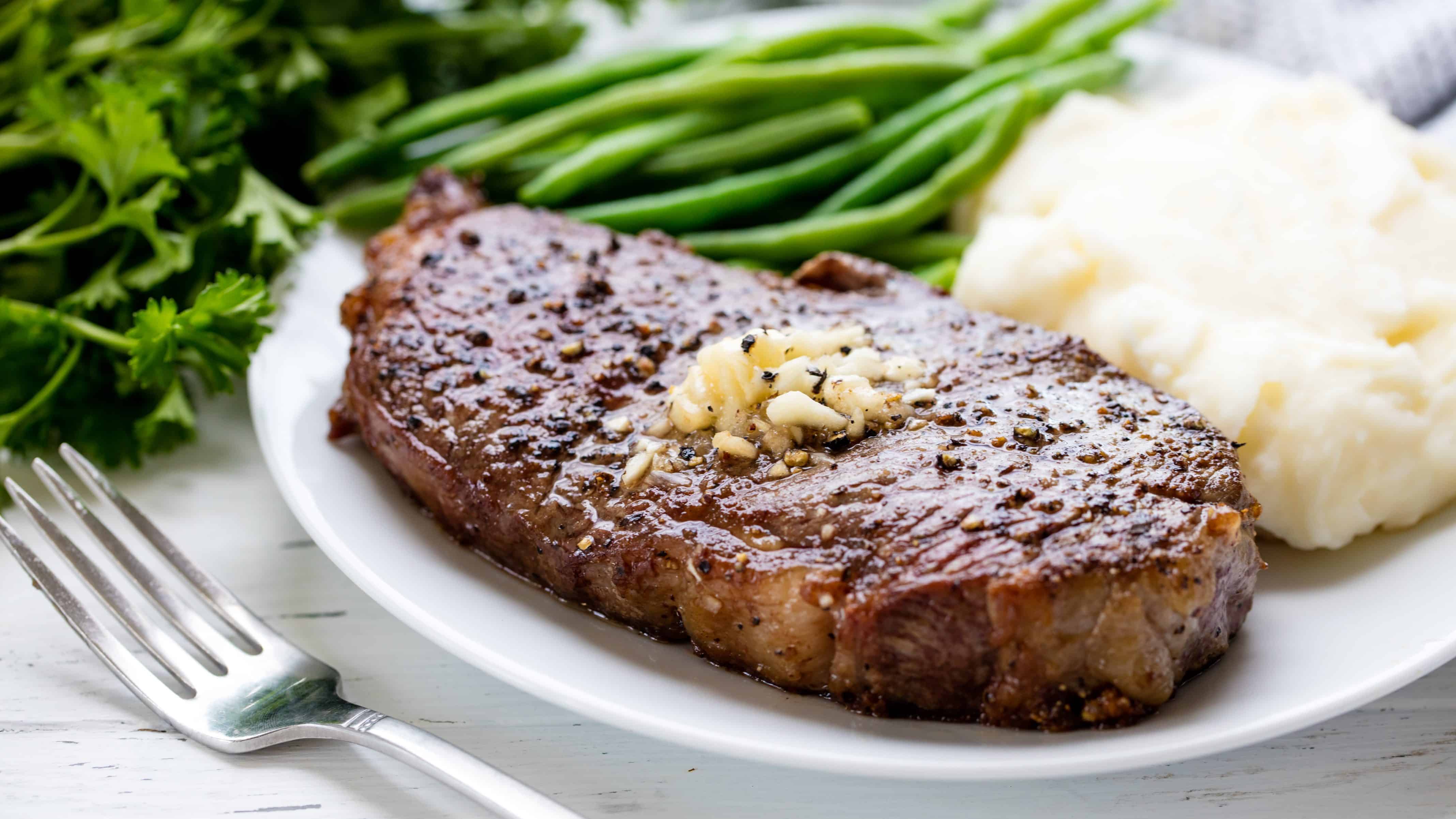 A juicy seasoned steak topped with a pat of butter and served with mashed potatoes and green beans on a plate.