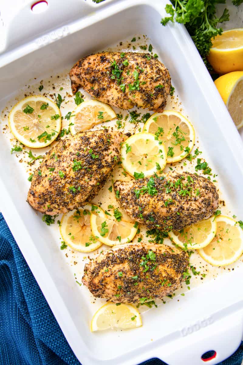 Baked chicken breasts in a pan seasoned with lemon pepper and chopped fresh parsley, surrounded by sliced lemon