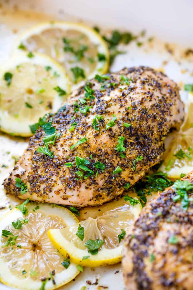 A baked chicken breast seasoned with lemon pepper and chopped fresh parley, surrounded by sliced lemon