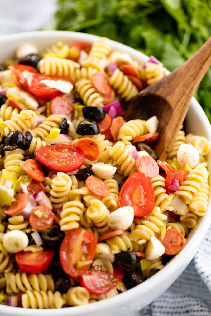A white bowl full of Pasta Salad with a large wooden spoon in it.