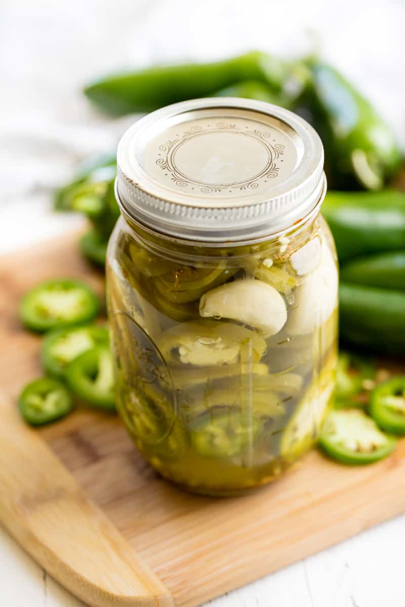 A mason jar filled with Pickled Jalapenos on a cutting board surrounded by fresh jalapenos