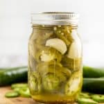 A mason jar filled with pickled jalapenos on a cutting board surrounded by fresh jalapenos