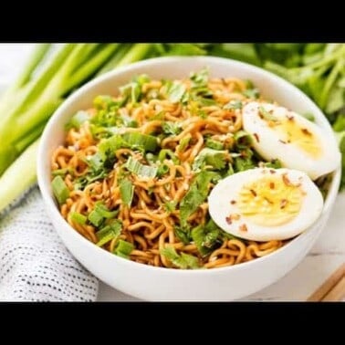 Ramen noodles in a white bowl topped with a boiled egg, green onions, and red pepper flakes.