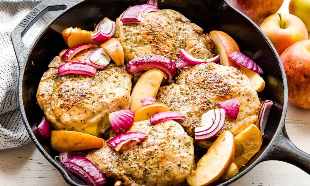 Apple stuffed pork chops in a skillet with onion slices and apple slices.