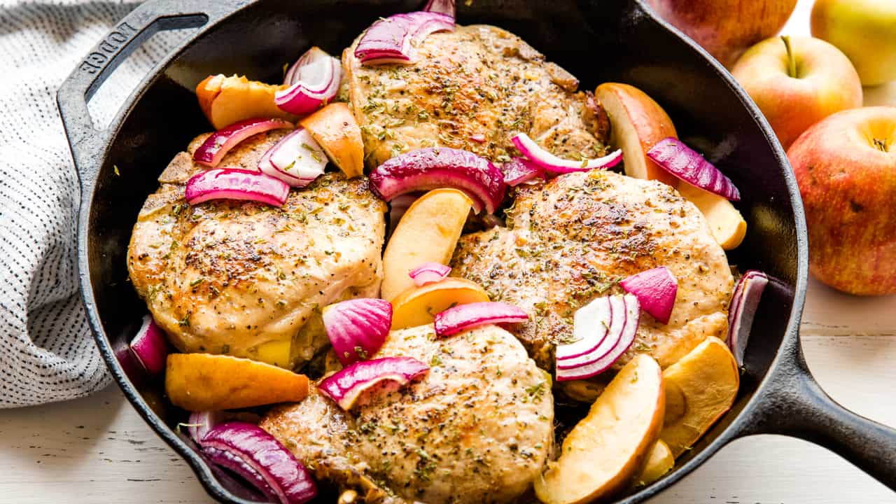 Apple stuffed pork chops in a skillet with onion slices and apple slices.