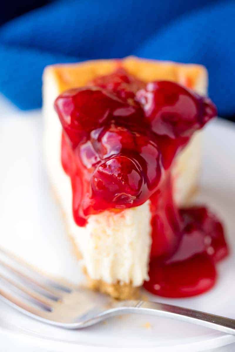Close up of a slice of New York Cheesecake topped a cherry topping with a bite taken out of it on a white plate.