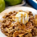 Apple Crisp served up on a white plate topped with a scoop of ice cream and caramel.