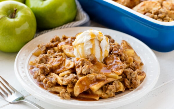 Apple crisp served up on a white plate topped with a scoop of ice cream and caramel sauce.