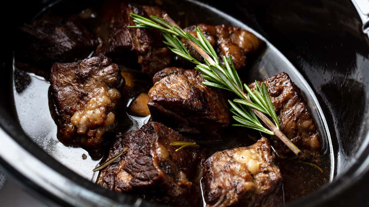 An overhead view of a slow cooker full of beef short ribs with a sprig of rosemary on top.