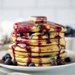 A stack of blueberry pancakes with a pad of butter on top and covered in blueberry syrup all on a white plate.