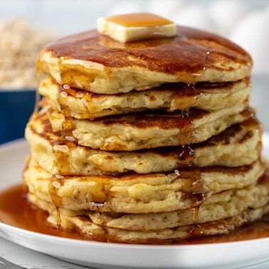 A stack of Oatmeal Pancakes topped with a pad of butter with syrup poured on top drizzling down the sides.