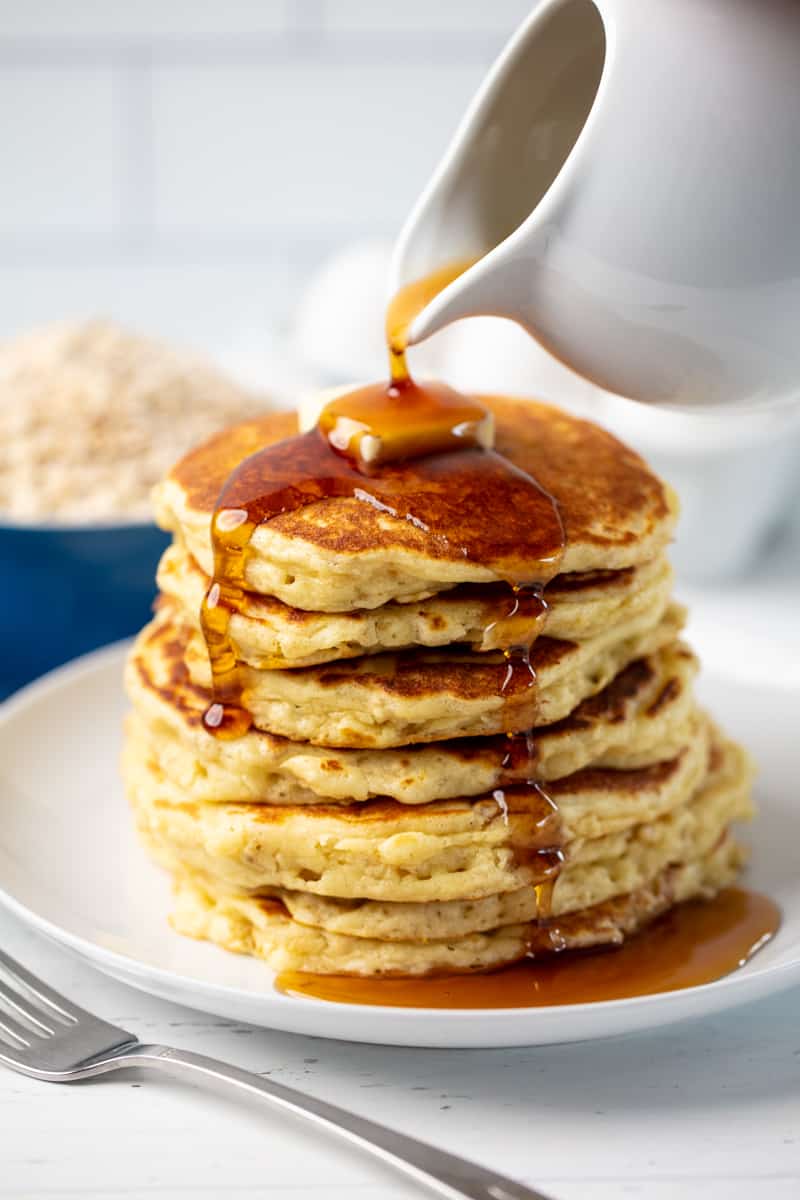 A stack of Oatmeal Pancakes topped with a pad of butter with syrup being poured on it.