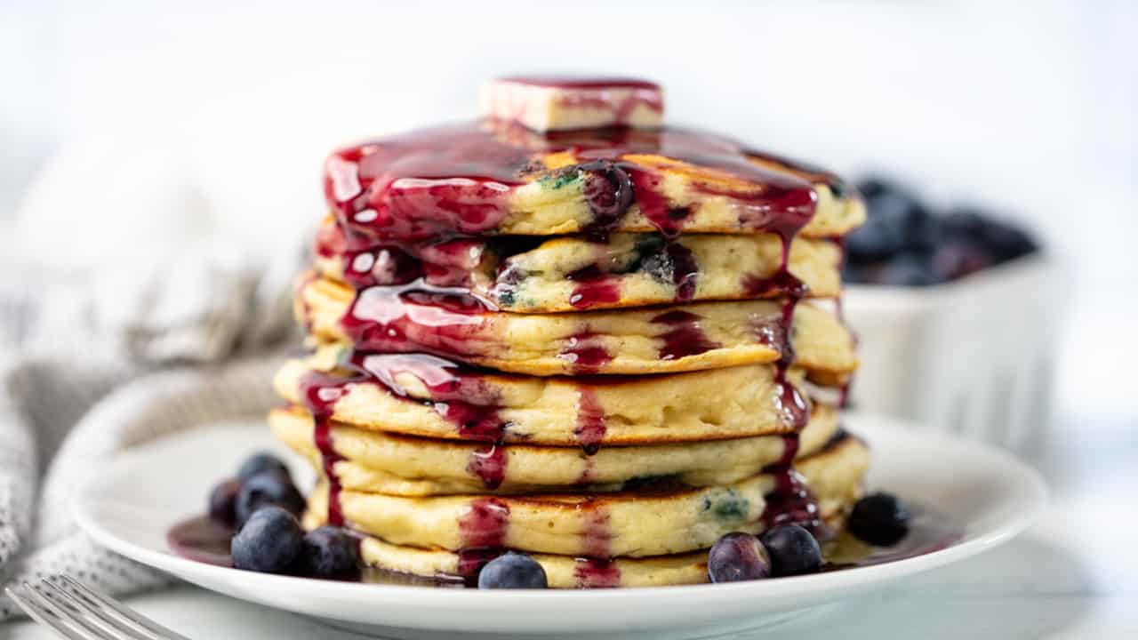 Pancakes stacked 7 high on a white plate with melted butter and blueberry syrup drizzled from top to bottom