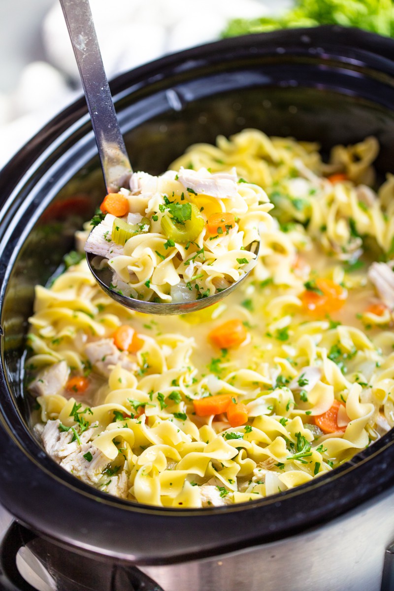 Ladle full of chicken noodle soup above a slow cooker full of chicken noodle soup.