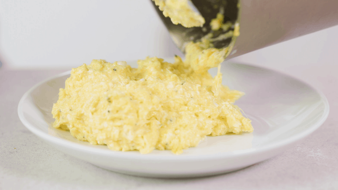 Scrambled eggs being poured out of a saucepan onto a white plate