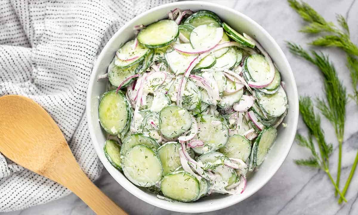 Bird's eye view of creamy cucumber salad in a white bowl with a wooden spoon next to it.