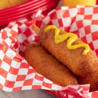 Three Corn Dogs in a basket with the top one topped with mustard.