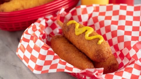 Three Corn Dogs in a basket with the top one topped with mustard.