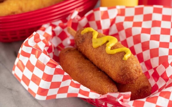 Three Corn Dogs in a basket with the top one topped with mustard.