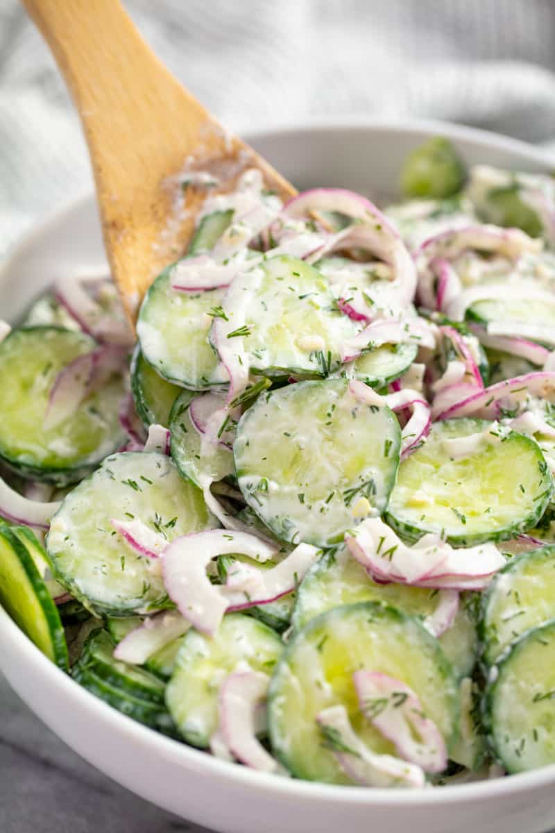 Close up of Creamy Cucumber Salad in a white bowl with a wooden spoon in it.