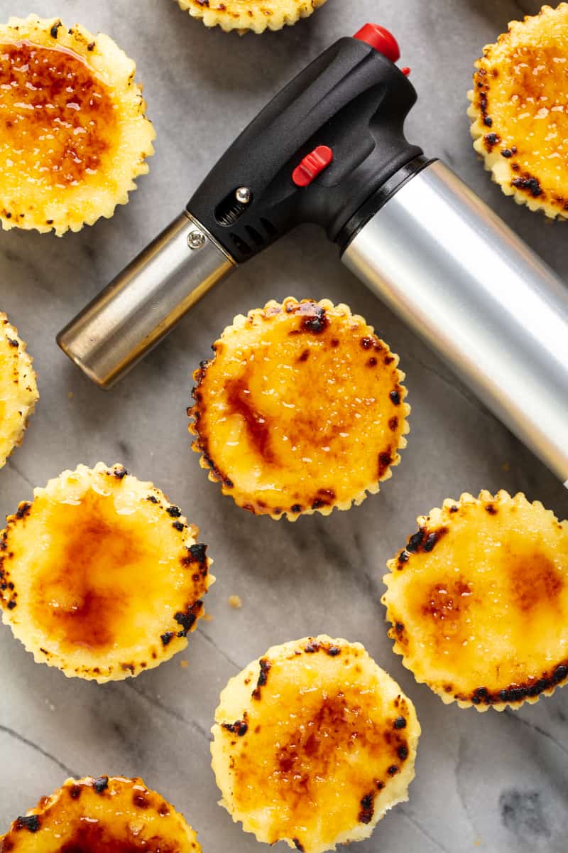 Bird's eye view of Creme Brulee Cheesecake Cupcakes and a kitchen torch on a marble countertop.