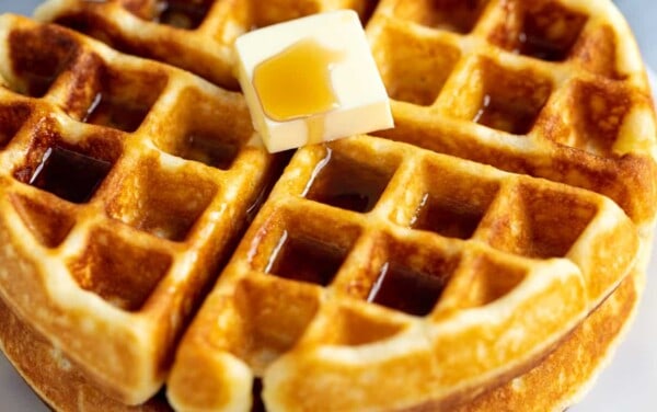 Stack of two waffles with a pad of butter and covered in syrup on a white plate.