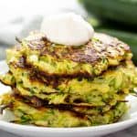 Stack of Zucchini Fritters topped with sour cream on a white plate.