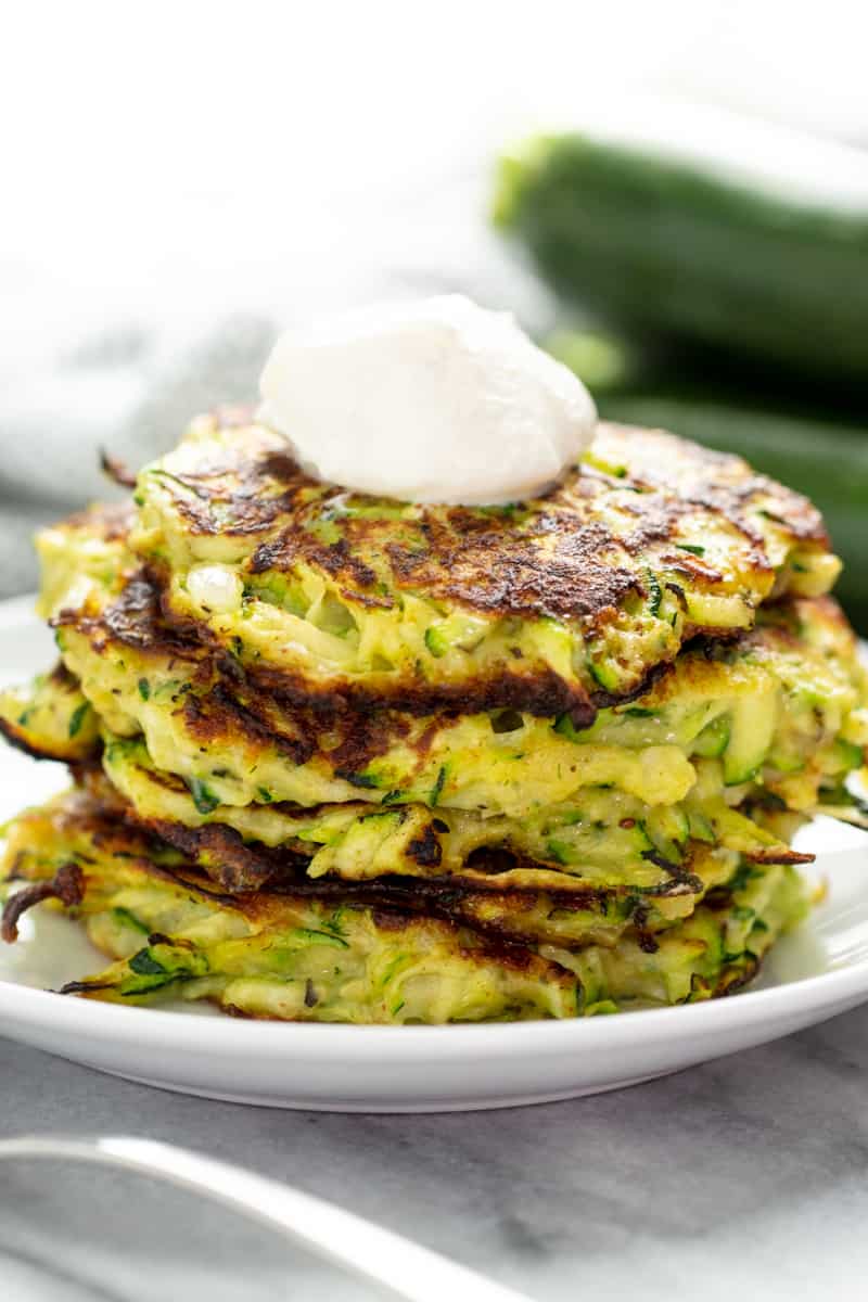 Stack of Zucchini Fritters topped with sour cream on a white plate.