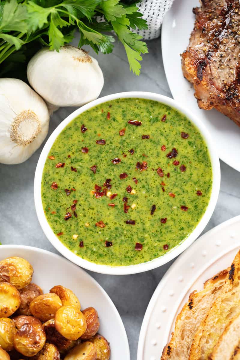 Bird's eye view of Chimichurri sauce in a white bowl surrounded by some other foods on the table.