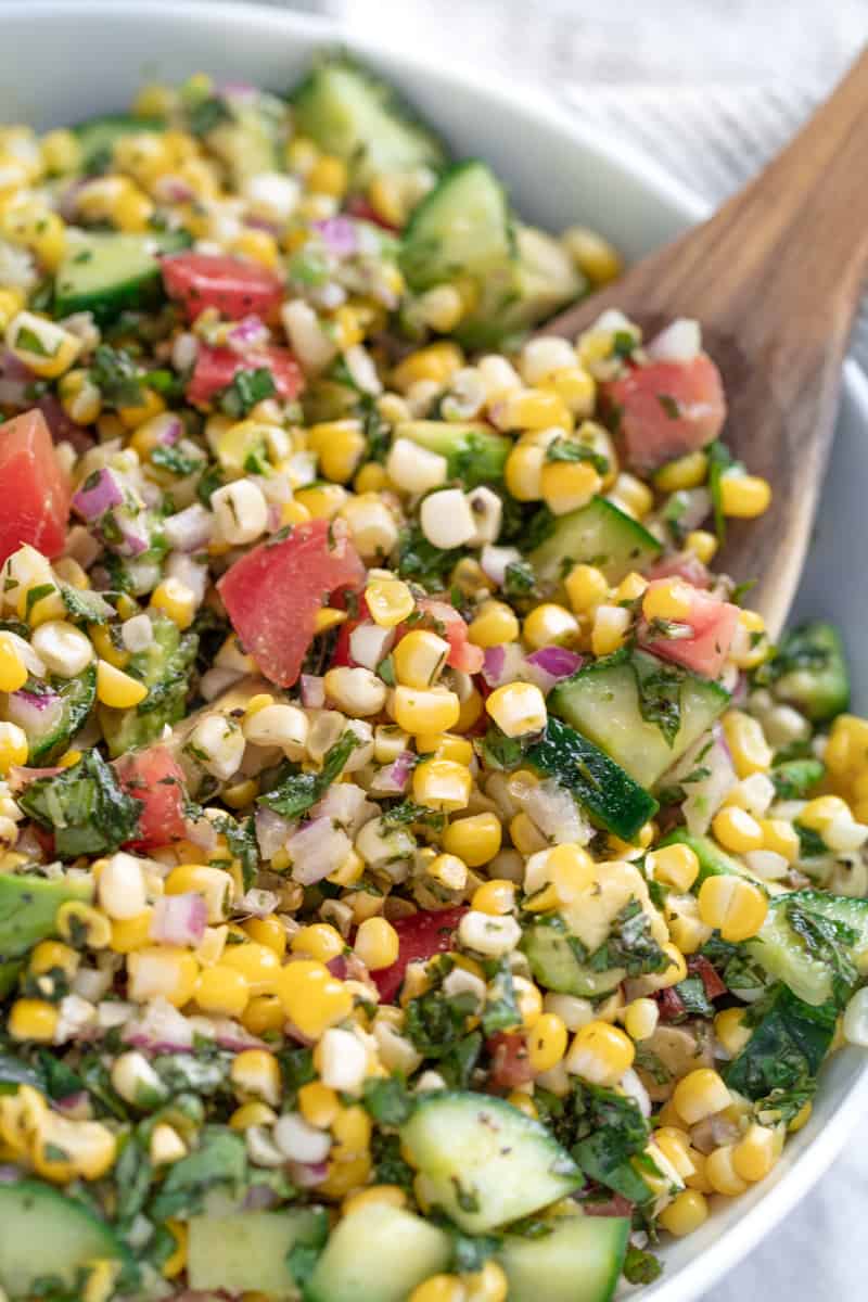 Corn salad in a white bowl with a wooden spoon in it.